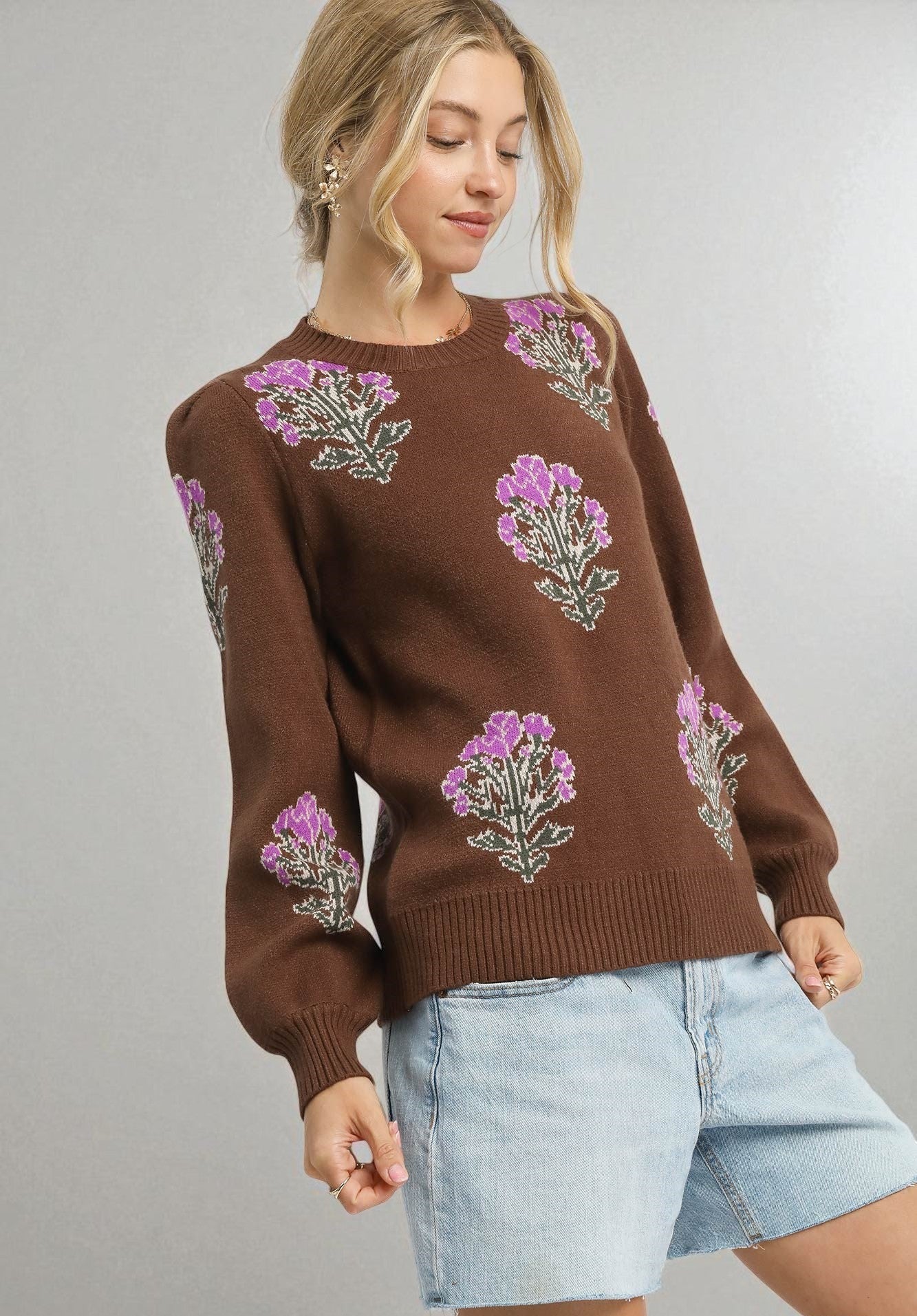 Woman wearing a brown sweater with floral patterns and denim shorts on a gray background