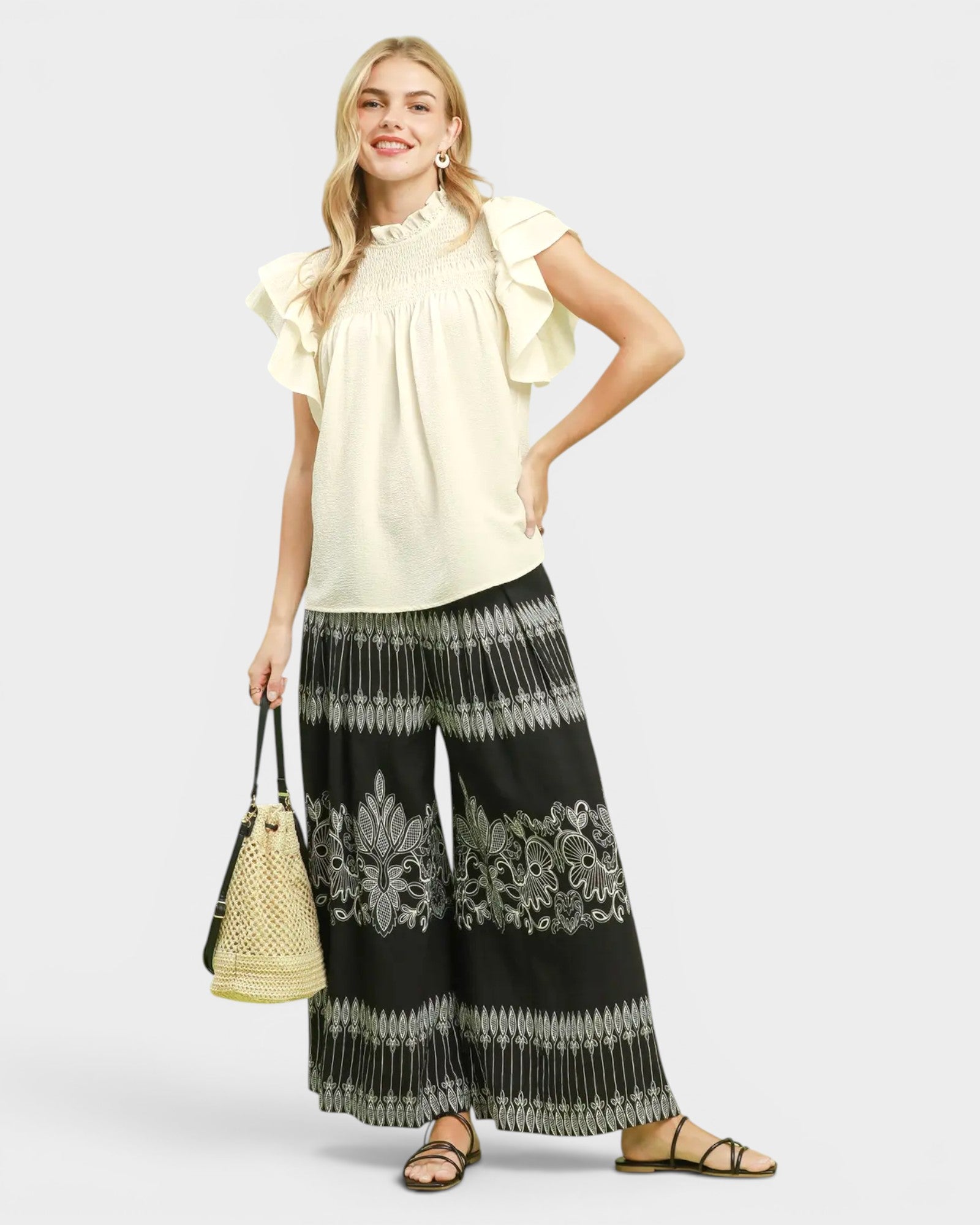 Woman wearing a cream blouse and black patterned pants, holding a beige handbag on a white background
