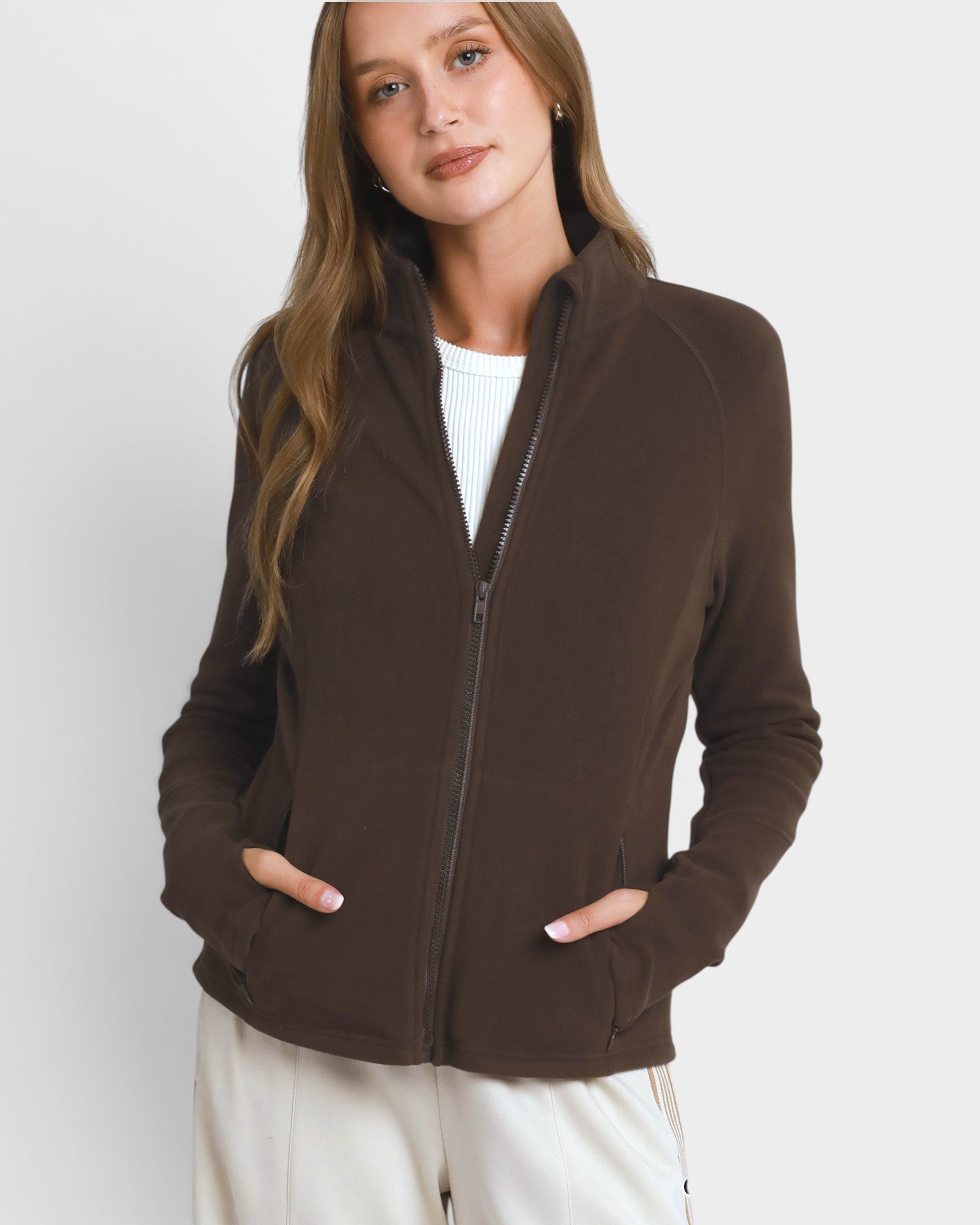 A woman wearing a dark brown zip-up fleece jacket over a white ribbed top and cream-colored pants, photographed against a light gray studio background.