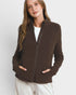 A woman wearing a dark brown zip-up fleece jacket over a white ribbed top and cream-colored pants, photographed against a light gray studio background.