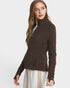A woman modeling a dark brown zip-up jacket with a mock neck and side pockets, paired with white pants featuring gold stripes, against a light gray studio background.