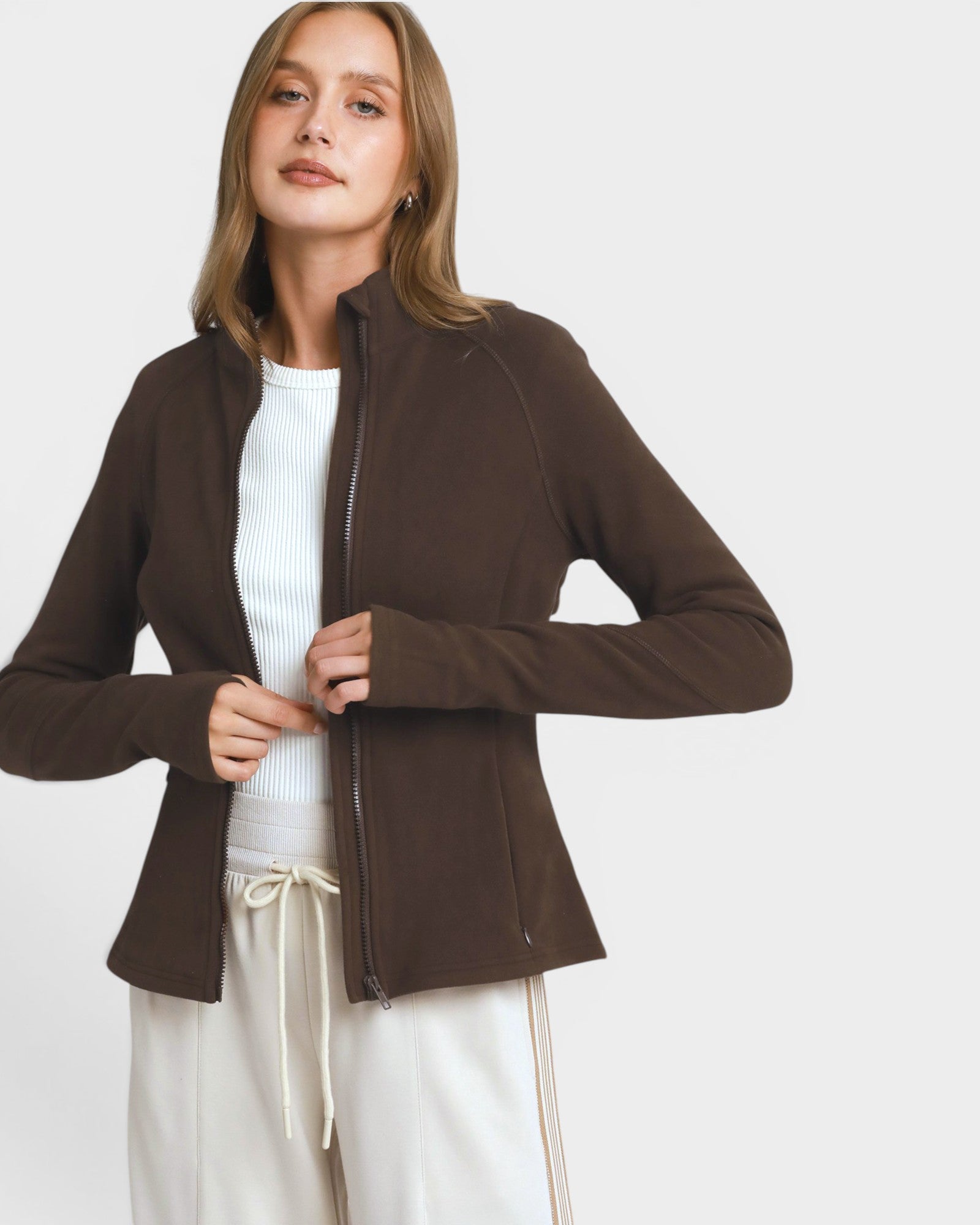 A woman wearing a fitted brown zip-front jacket over a white ribbed top and white drawstring pants, photographed against a light gray studio background.