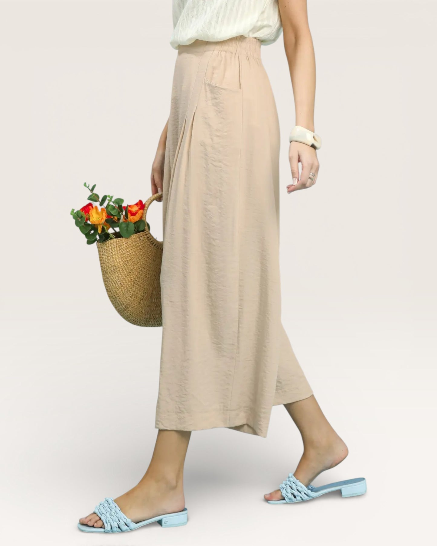 Person wearing a beige skirt holding a woven bag with flowers on a plain background
