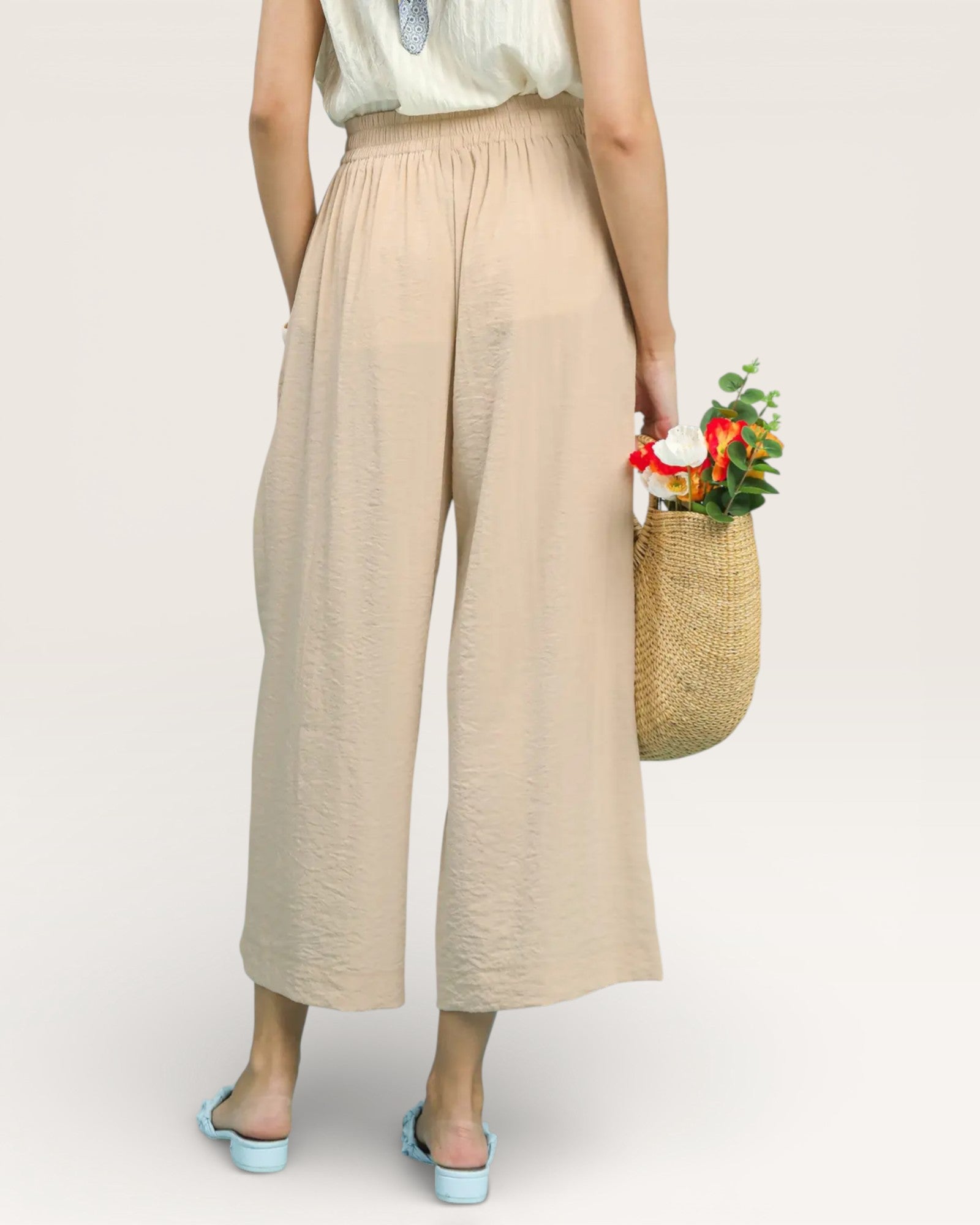 Person wearing beige wide-leg pants holding a woven bag with flowers on a plain background