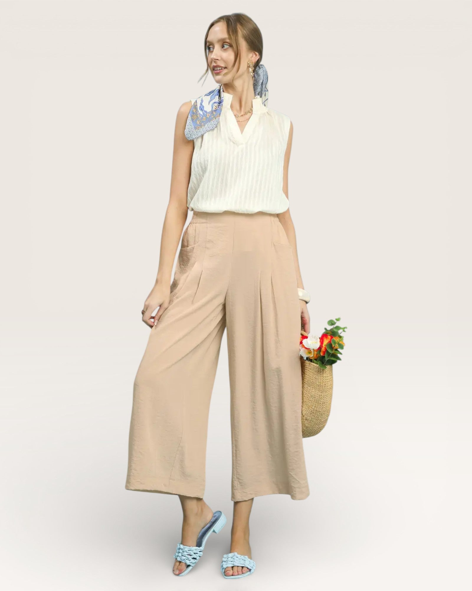 Woman wearing a white sleeveless top and beige wide-leg pants, holding flowers and a basket, on a light gray background.