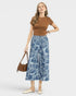 Woman wearing a brown top and blue patterned pants with a handbag on a white background