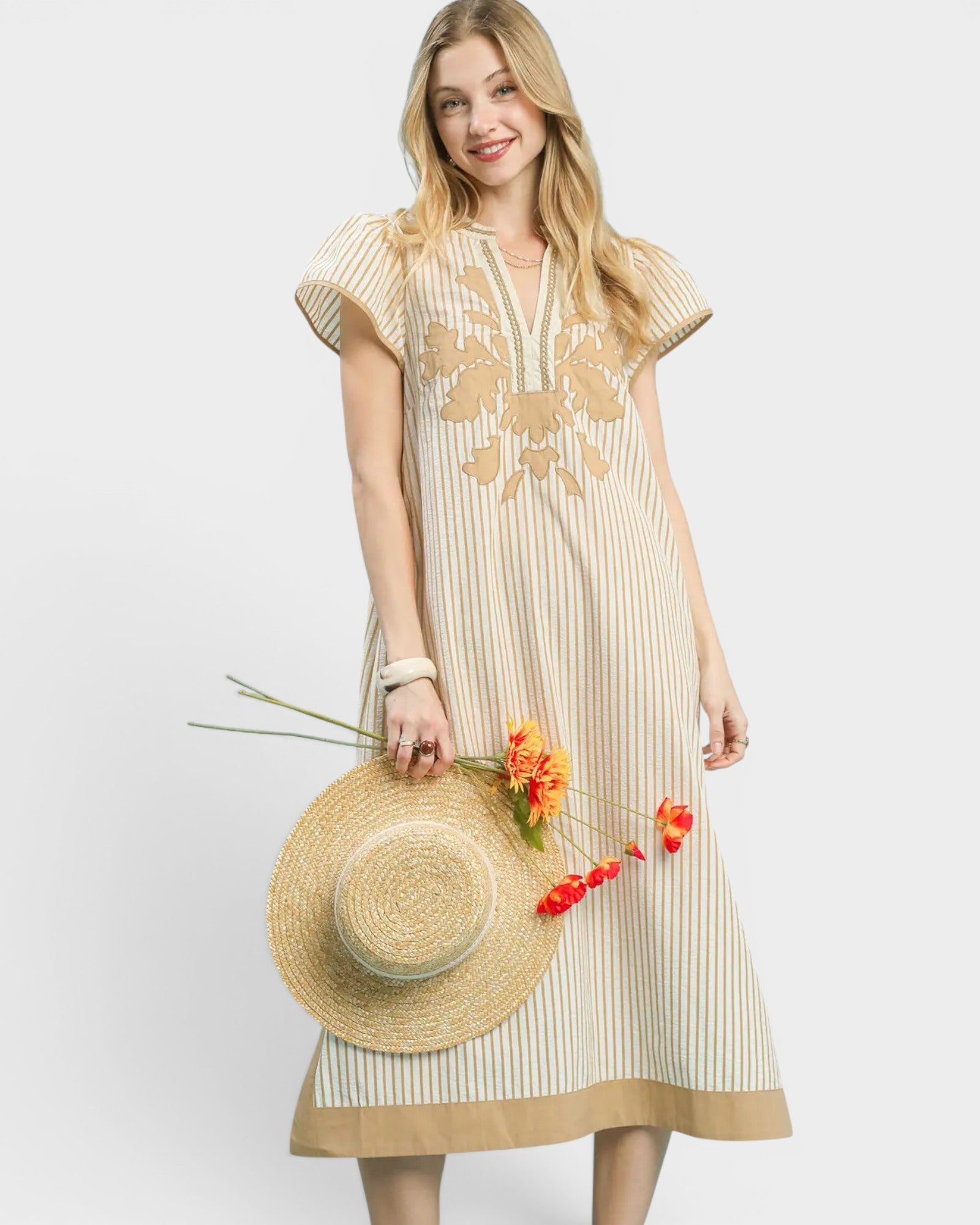 Woman wearing a beige dress with floral patterns, holding a straw hat and flowers on a white background