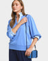 Woman wearing a blue blouse with rolled-up sleeves, holding a matching blue handbag against a light background.