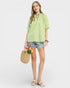 Woman wearing a green blouse and denim shorts, holding flowers and a straw bag on a white background