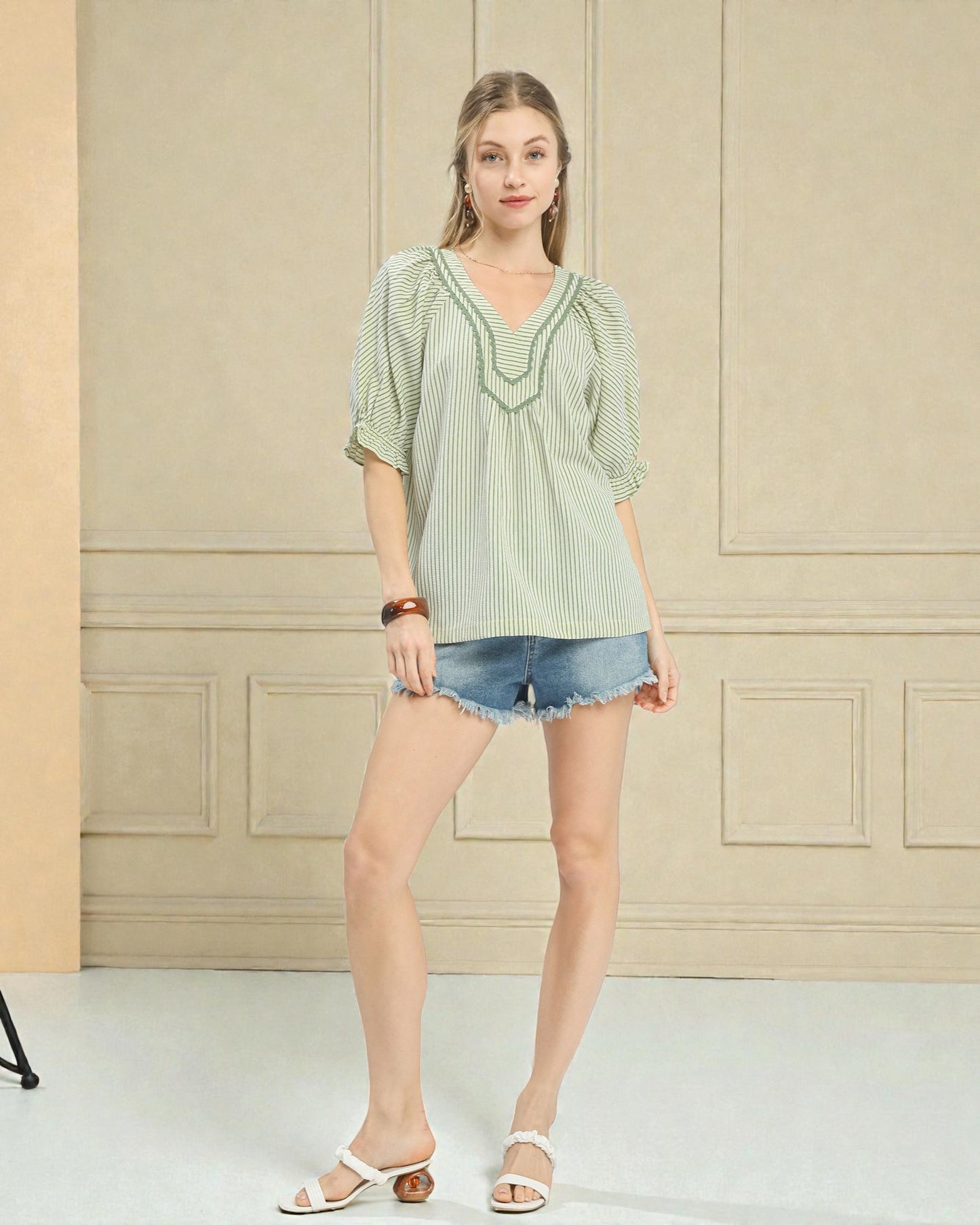 Woman wearing a light green striped blouse and denim shorts standing against a neutral wall.
