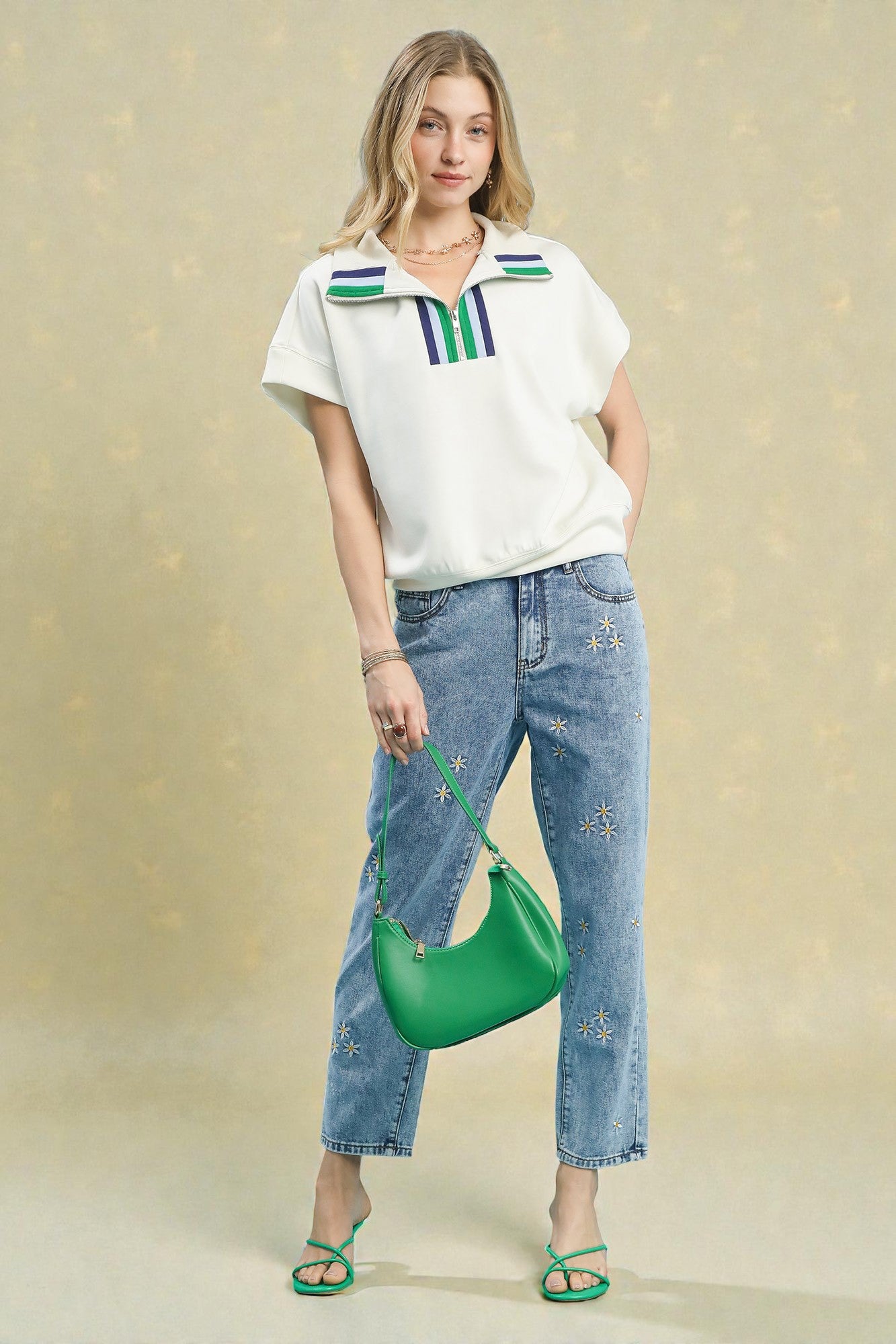Woman wearing a white shirt with colorful accents and blue jeans, holding a green handbag against a beige background.