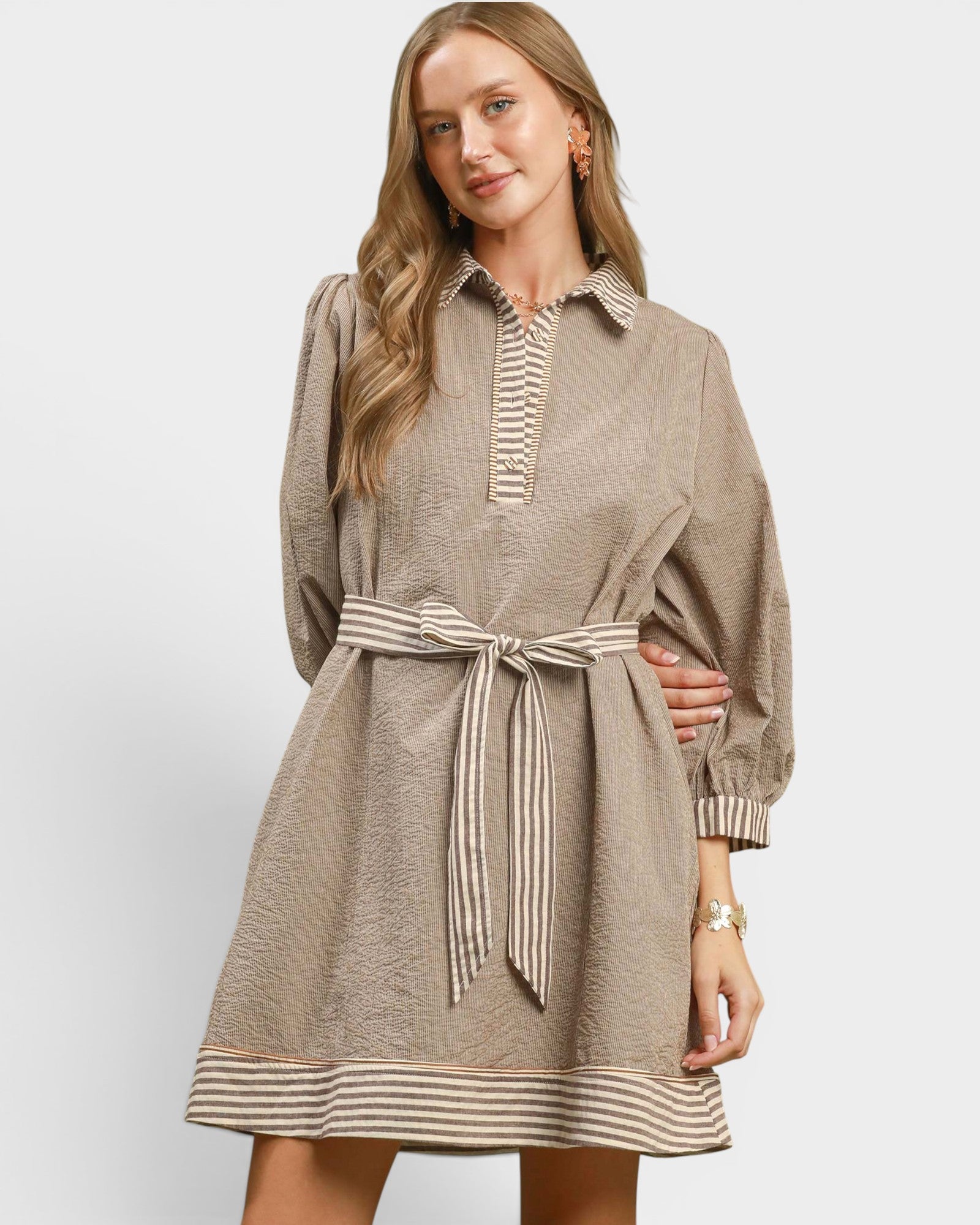 A woman modeling a taupe-brown long-sleeve mini dress with a striped collar, button placket, cuffs, hem, and matching tie belt, set against a plain light gray studio background.