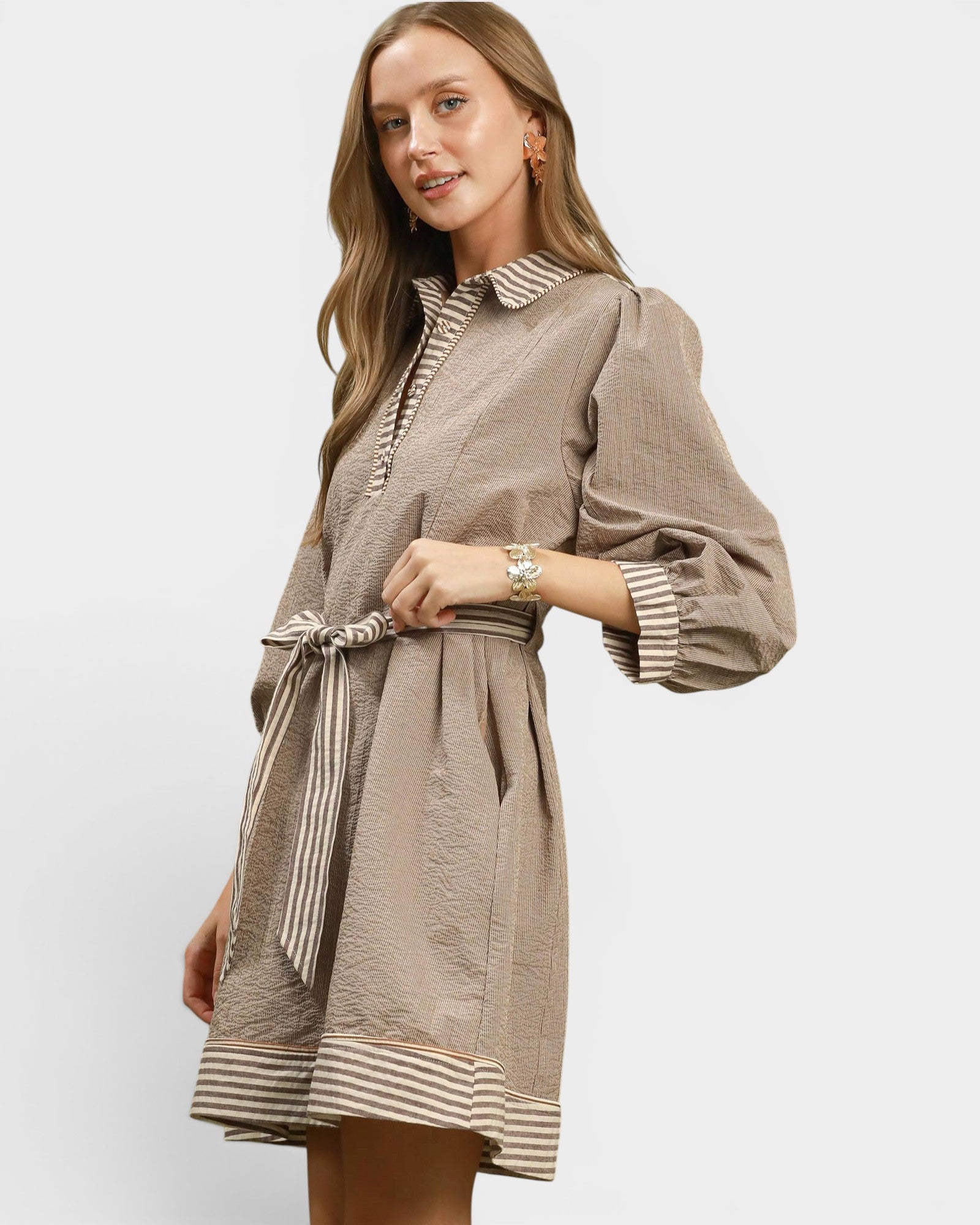 A woman modeling a taupe-brown mini dress with a striped collar, cuffs, hem, and matching tie belt, set against a plain light gray studio background.