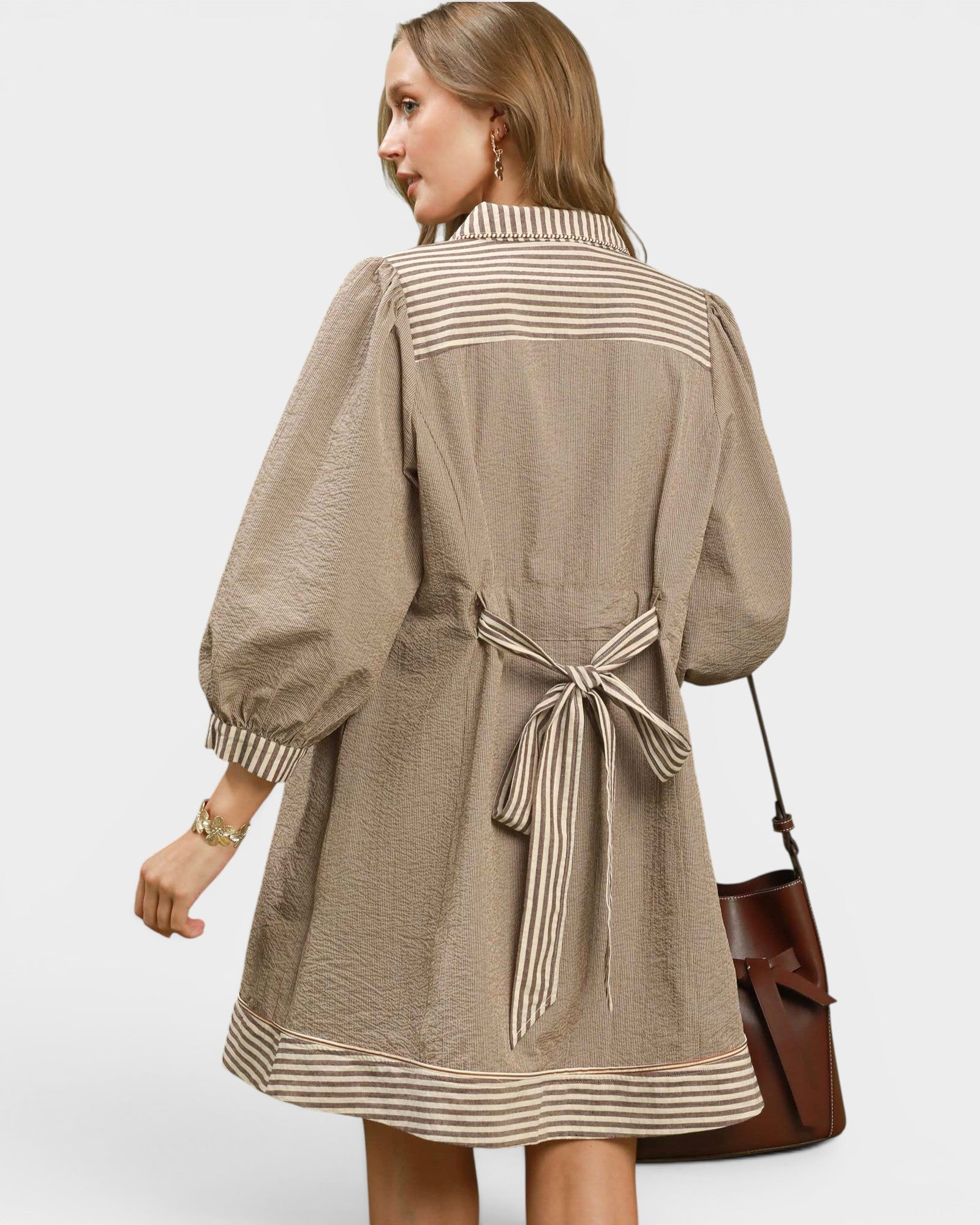 A woman shown from the back wearing a beige and white striped long-sleeve tunic dress with a tie belt at the waist, paired with a brown leather shoulder bag, against a light gray studio background.