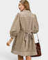 A woman shown from the back wearing a beige and white striped long-sleeve tunic dress with a tie belt at the waist, paired with a brown leather shoulder bag, against a light gray studio background.
