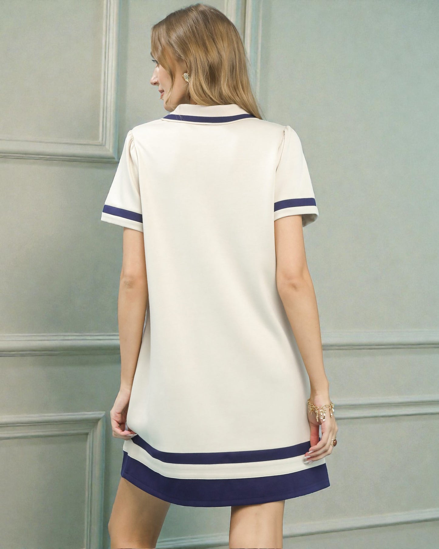 Woman wearing a white dress with blue trim against a light gray wall.