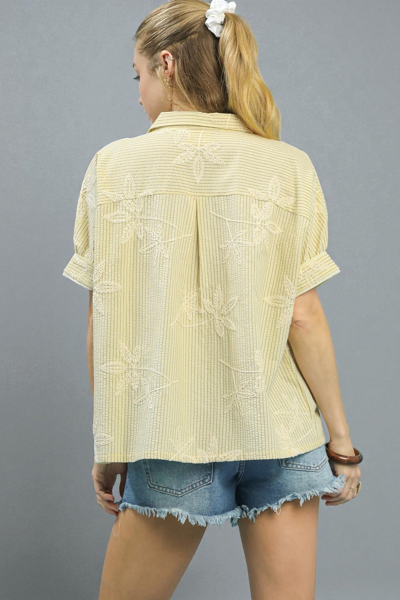 Woman wearing a light yellow textured shirt and denim shorts on a gray background