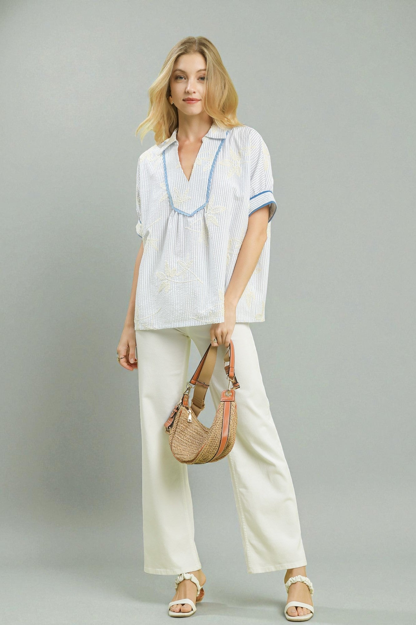 Woman wearing a white outfit with a blue trim, holding a woven bag on a gray background