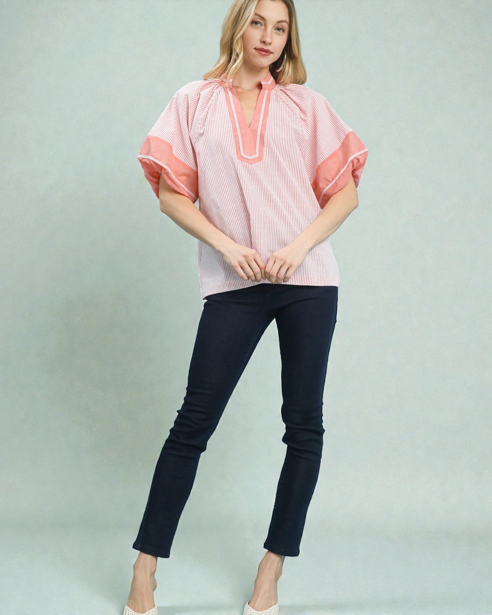Woman wearing a pink blouse with white trim and black pants on a light gray background
