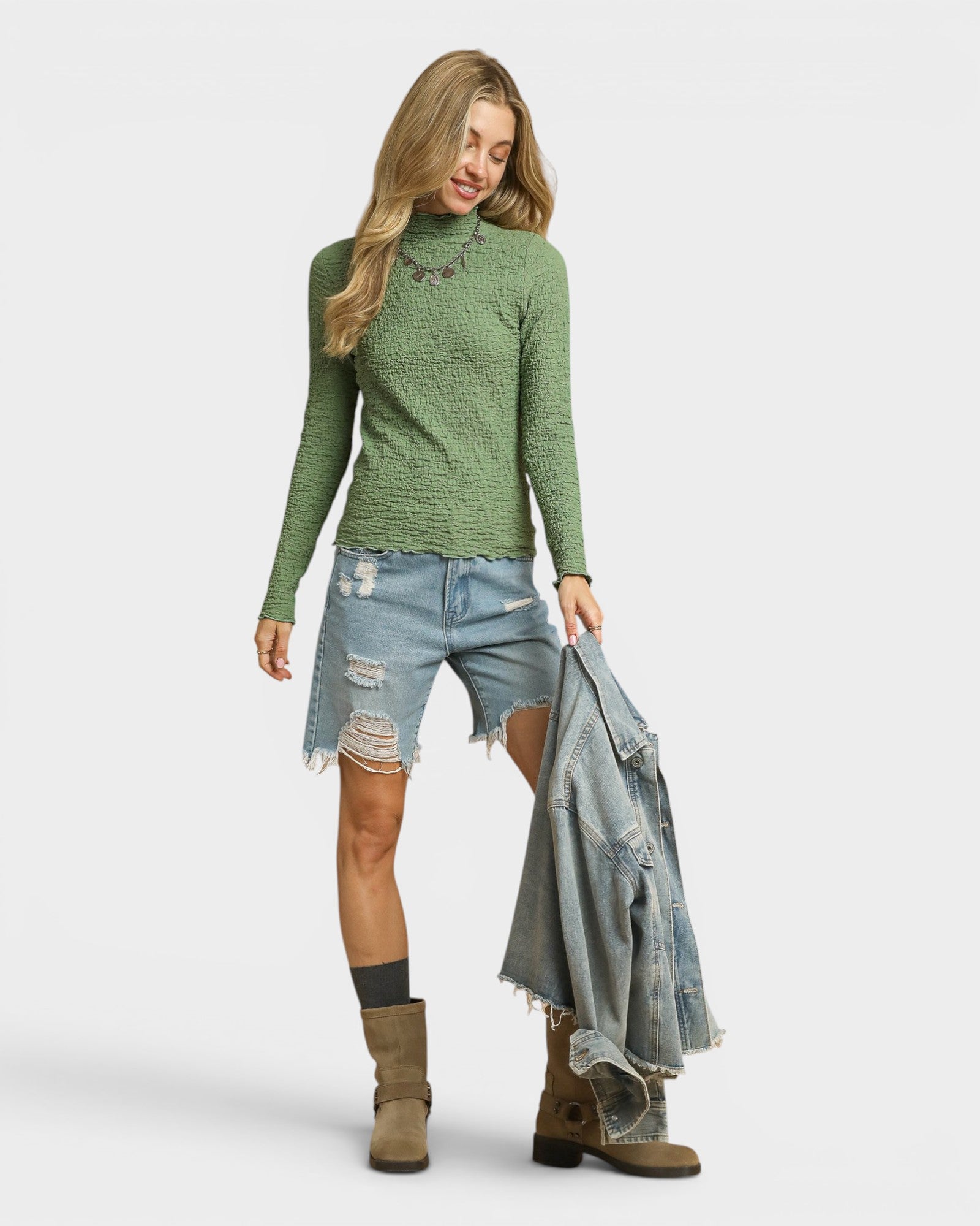 Woman wearing a green patterned sweater, denim shorts, and boots holding a denim jacket on a white background