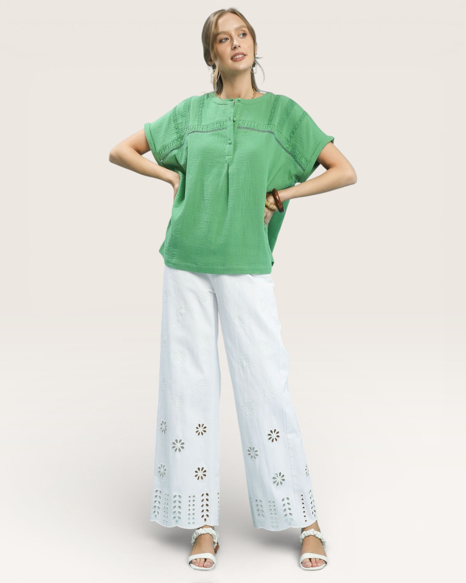 Woman wearing a green top and white pants with eyelet details on a beige background