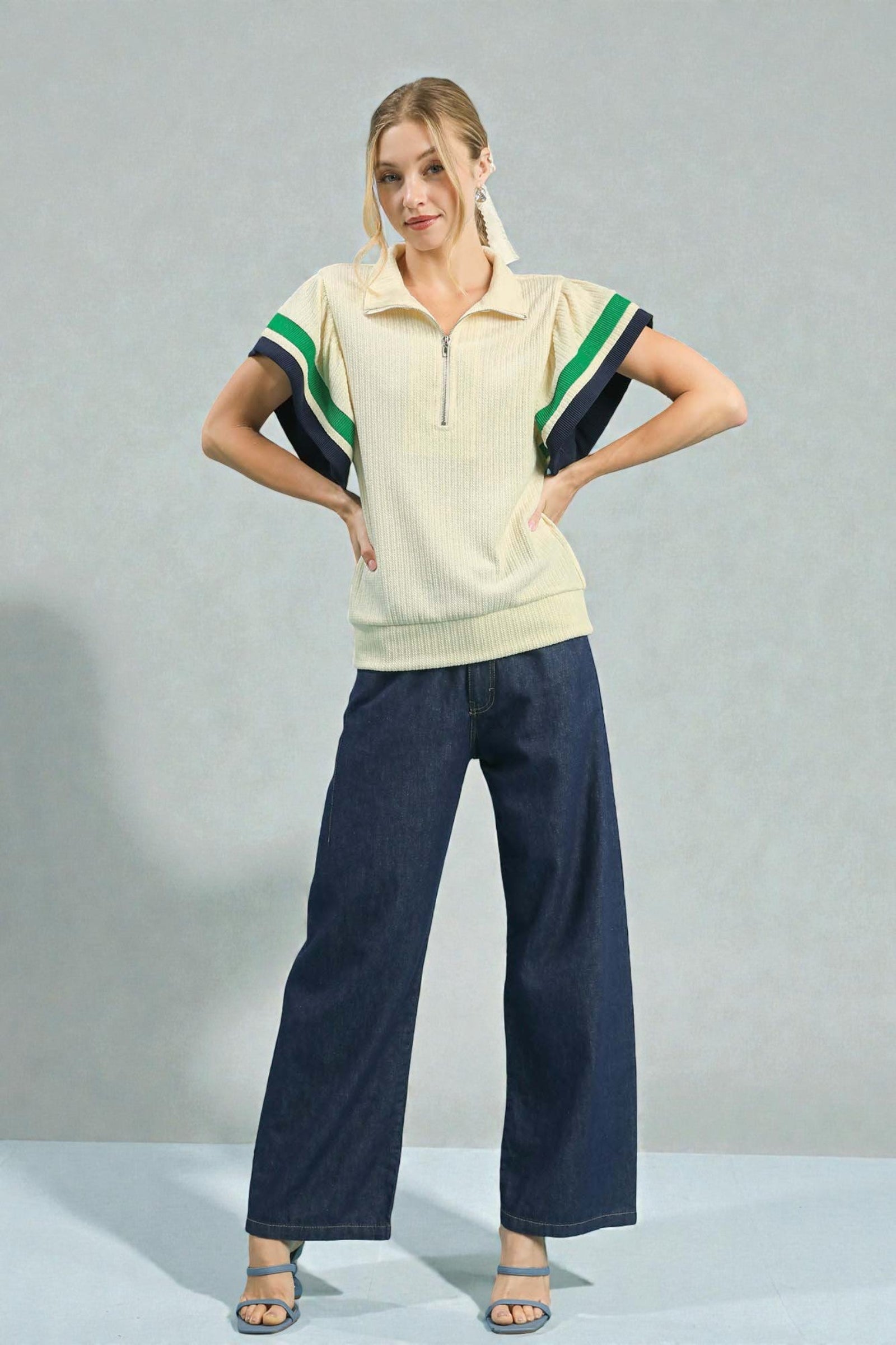 Woman wearing a cream-colored polo shirt with green and navy accents and dark blue pants on a gray background