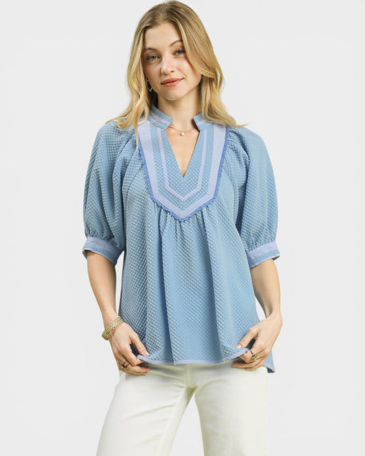 Woman wearing a blue patterned blouse with white pants on a light gray background