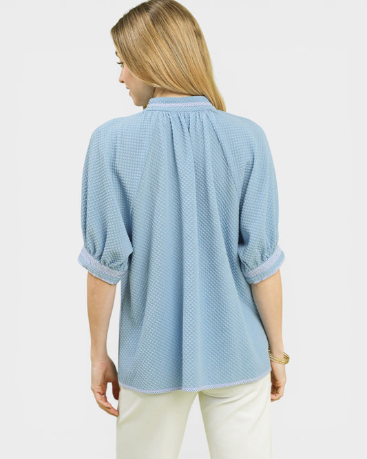 Woman wearing a light blue textured blouse with white pants on a plain background