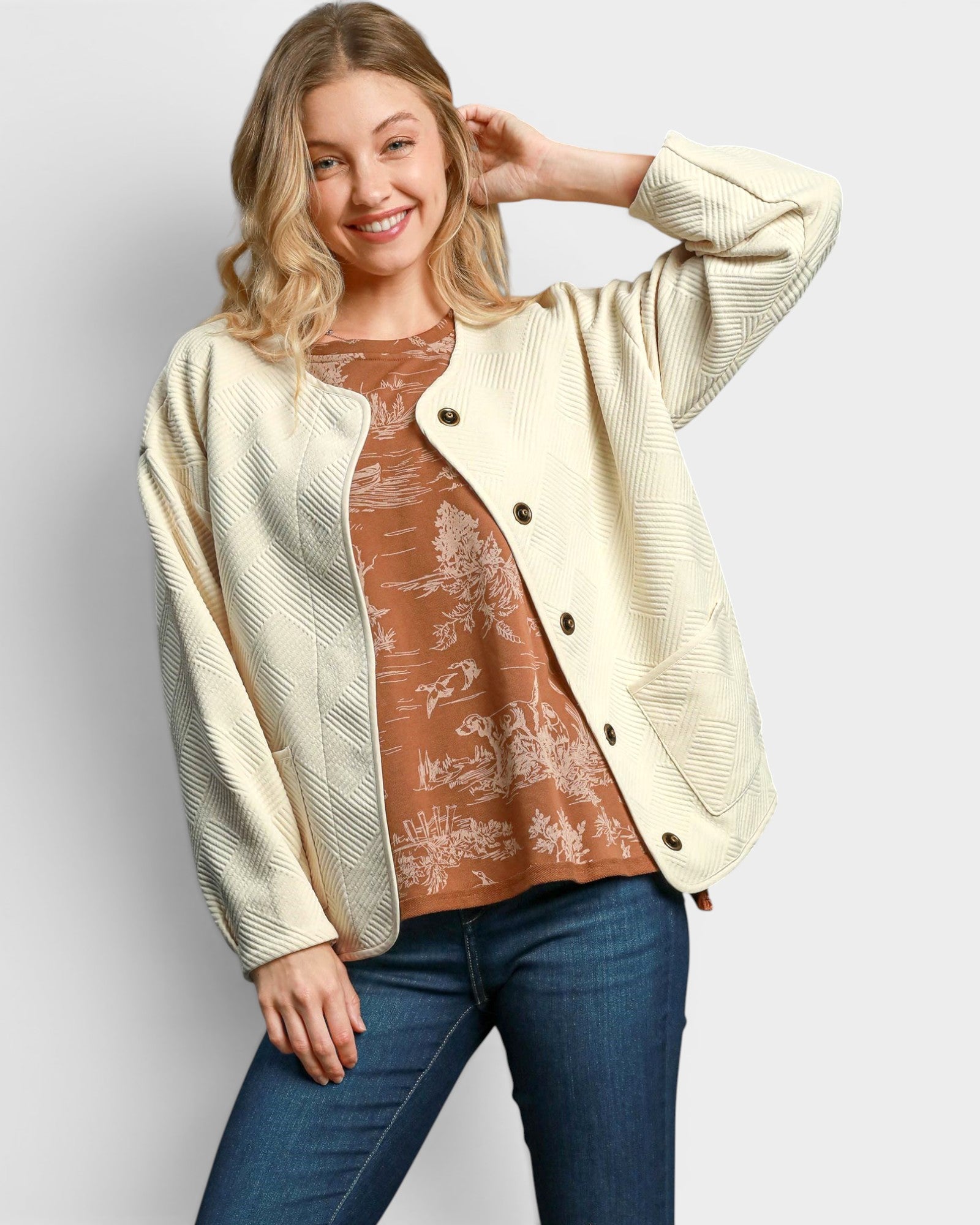 Woman wearing a cream cardigan over a brown patterned top and blue jeans on a light gray background