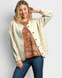 Woman wearing a cream cardigan over a brown patterned top and blue jeans on a light gray background