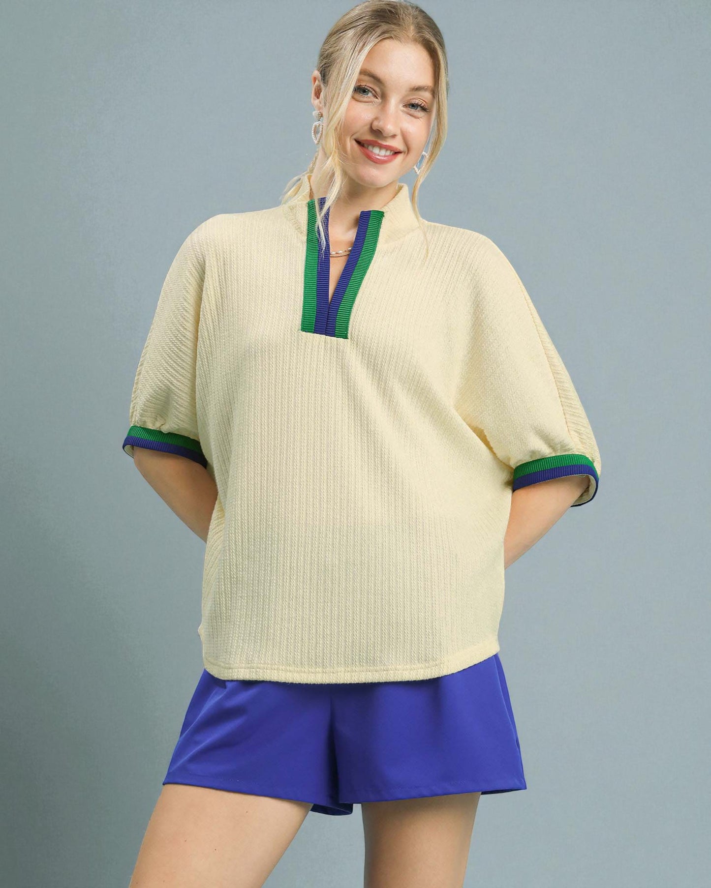 Woman wearing a beige top with green and blue trim and purple shorts on a gray background