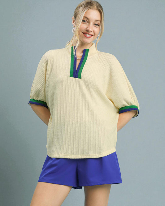 Woman wearing a beige top with green and blue trim and purple shorts on a gray background