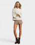 Woman wearing a white sweater and beige shorts with brown boots on a plain background