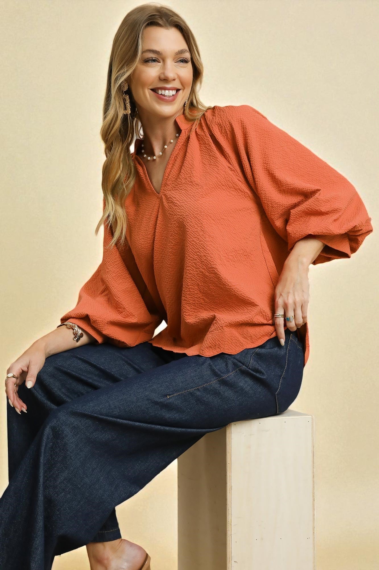 Woman wearing an orange blouse and navy pants sitting on a beige background