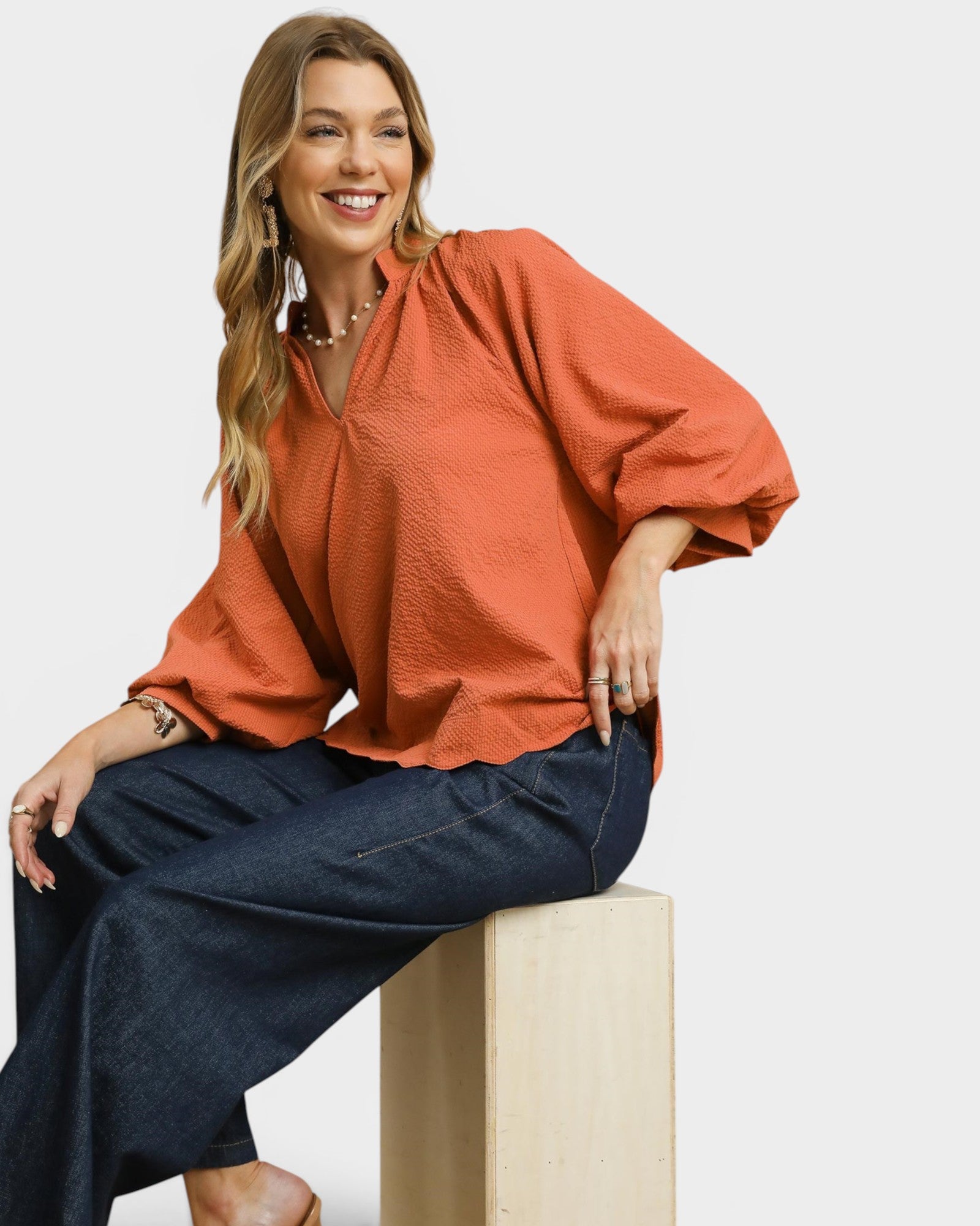 Woman wearing an orange blouse and navy pants sitting on a white background