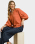 Woman wearing an orange blouse and navy pants sitting on a white background
