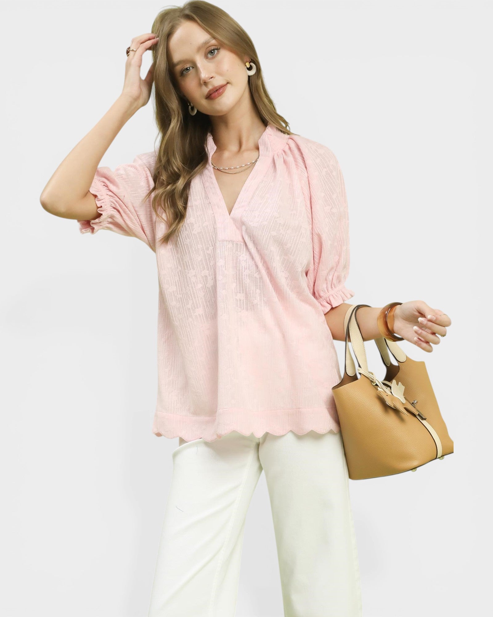 Woman wearing a pink blouse and white pants, holding a beige handbag on a light gray background