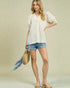 Woman wearing a white blouse and denim shorts, holding a straw bag with a blue scarf.