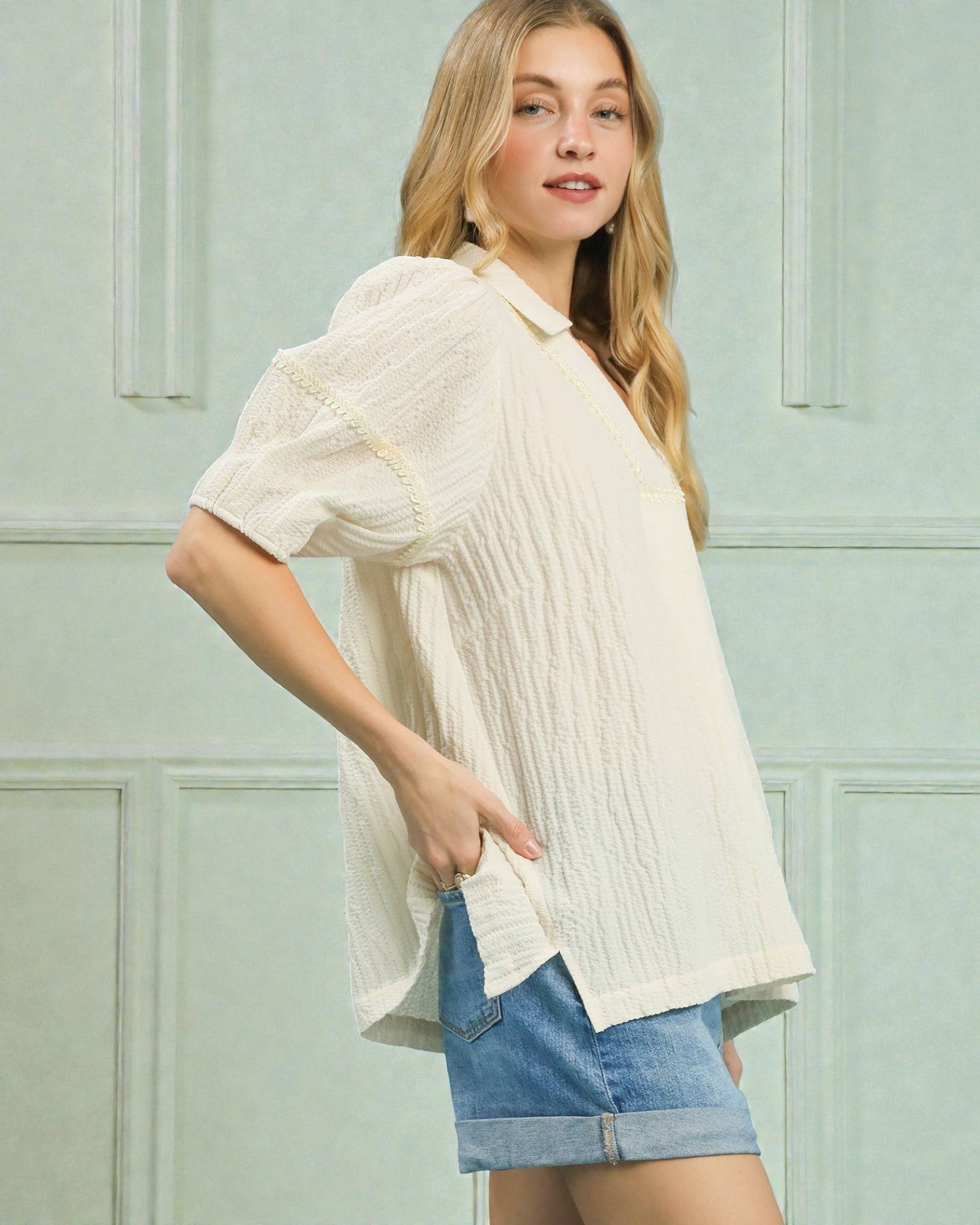 Woman wearing a white blouse and denim shorts against a light green wall.