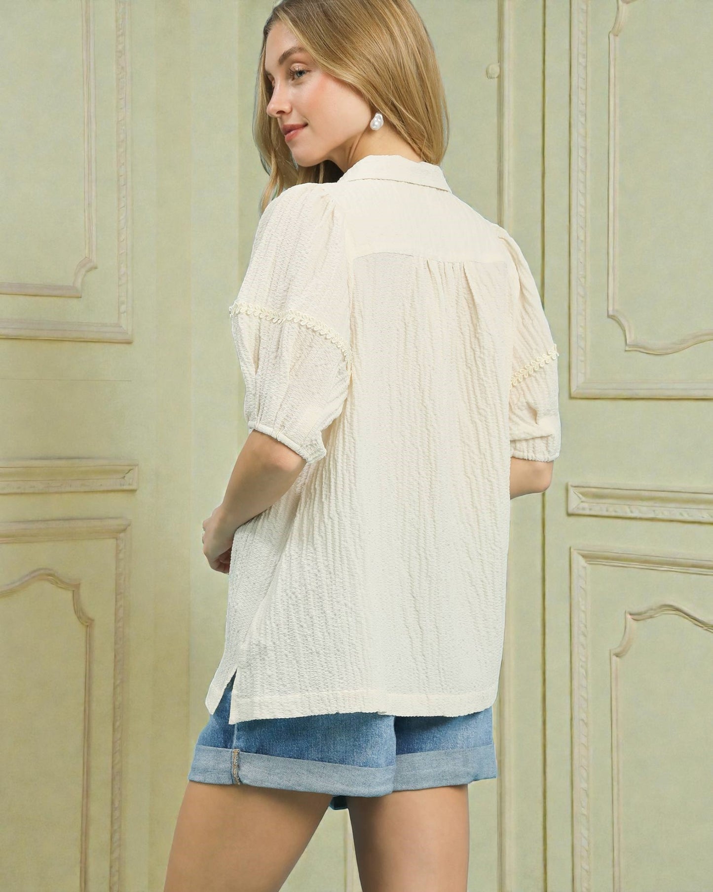 Woman wearing a white blouse and denim shorts standing in front of a light green wall.