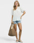 Woman wearing a light blue shirt and denim shorts, holding a woven bag on a white background
