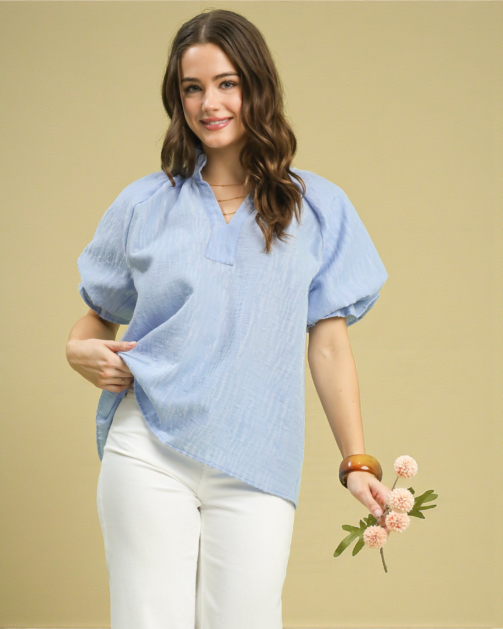 Woman wearing a light blue blouse and white pants, holding flowers against a beige background