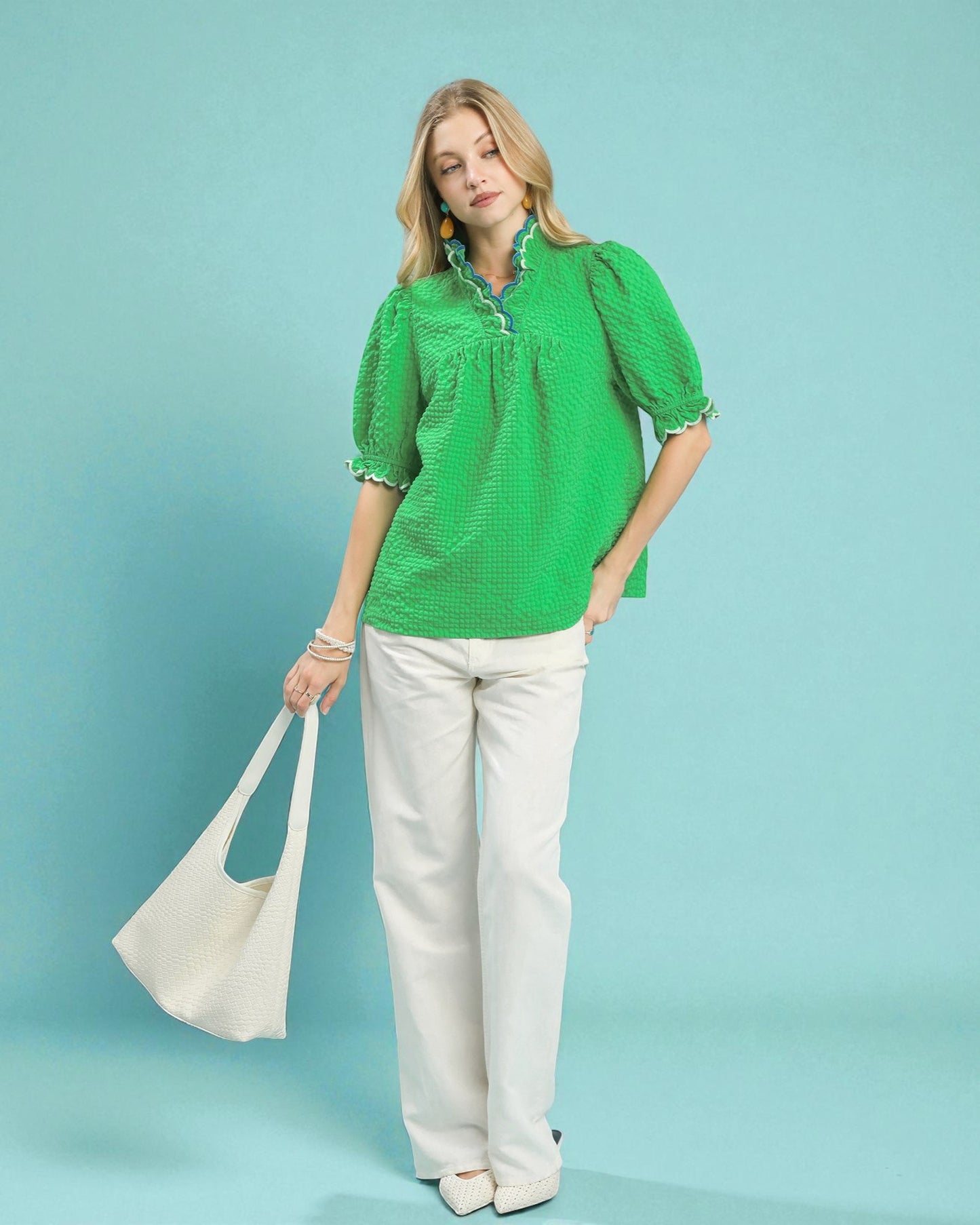 Woman wearing a green blouse and white pants holding a beige bag against a teal background