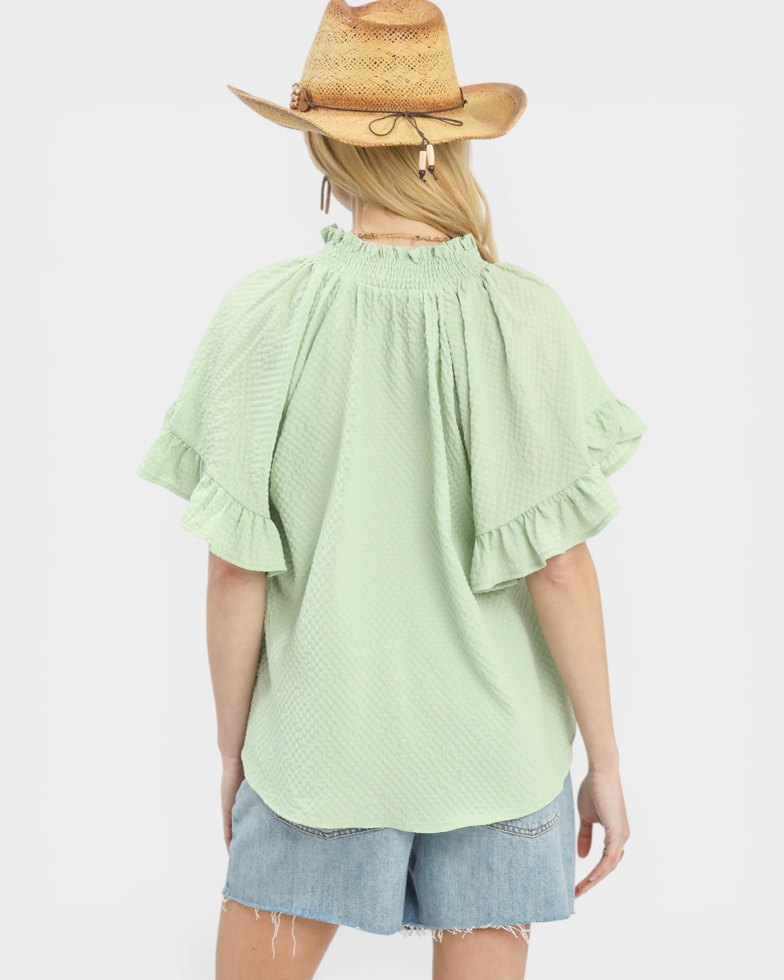 Person wearing a light green blouse with ruffled sleeves and denim shorts on a white background