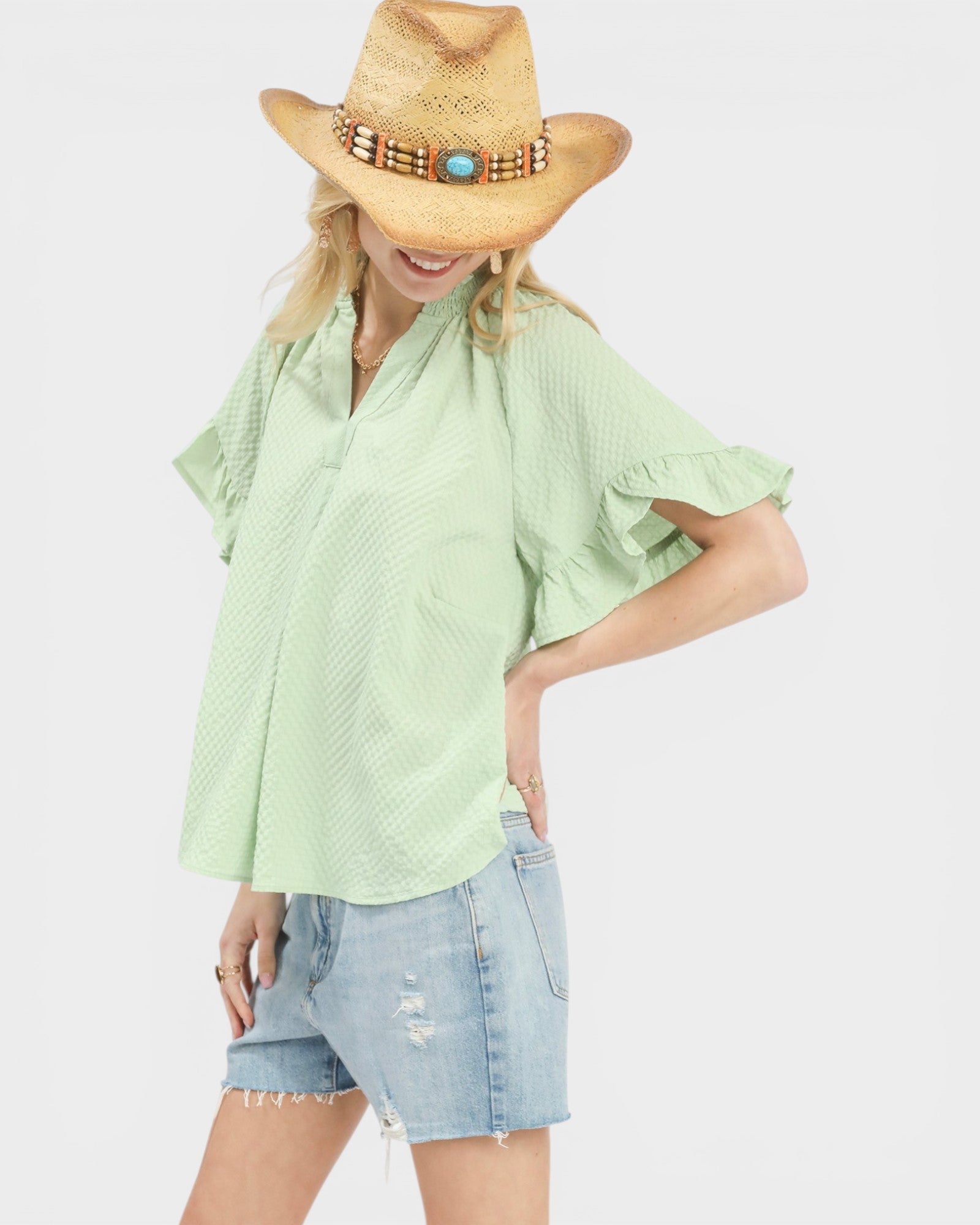 Woman wearing a light green blouse, denim shorts, and a straw hat with beads on a white background