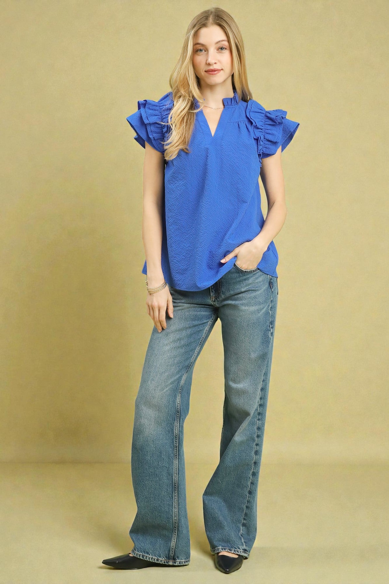 Woman wearing a blue blouse with ruffled sleeves and blue jeans on a beige background