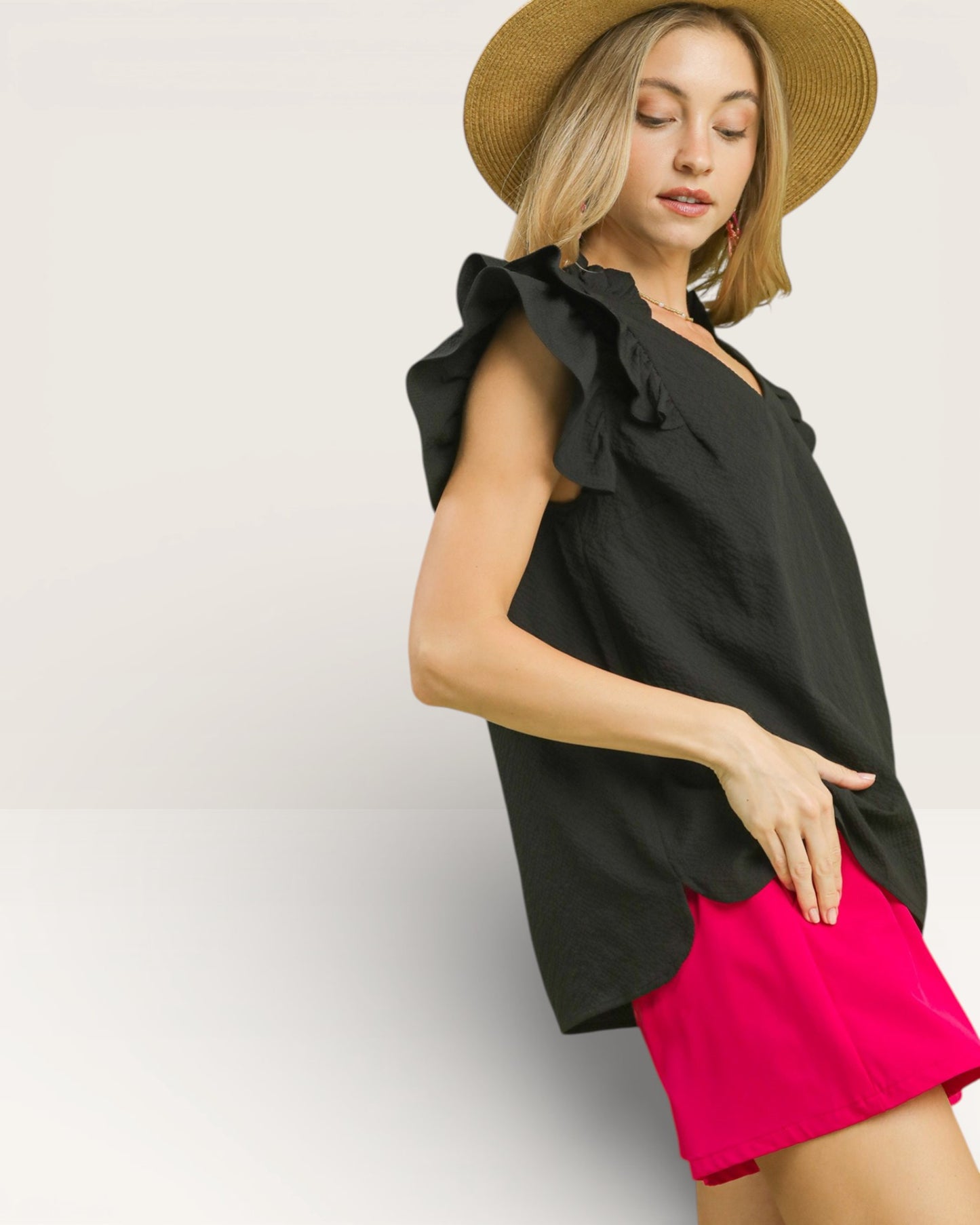 Woman wearing a black sleeveless top with ruffled shoulders and pink shorts on a light gray background