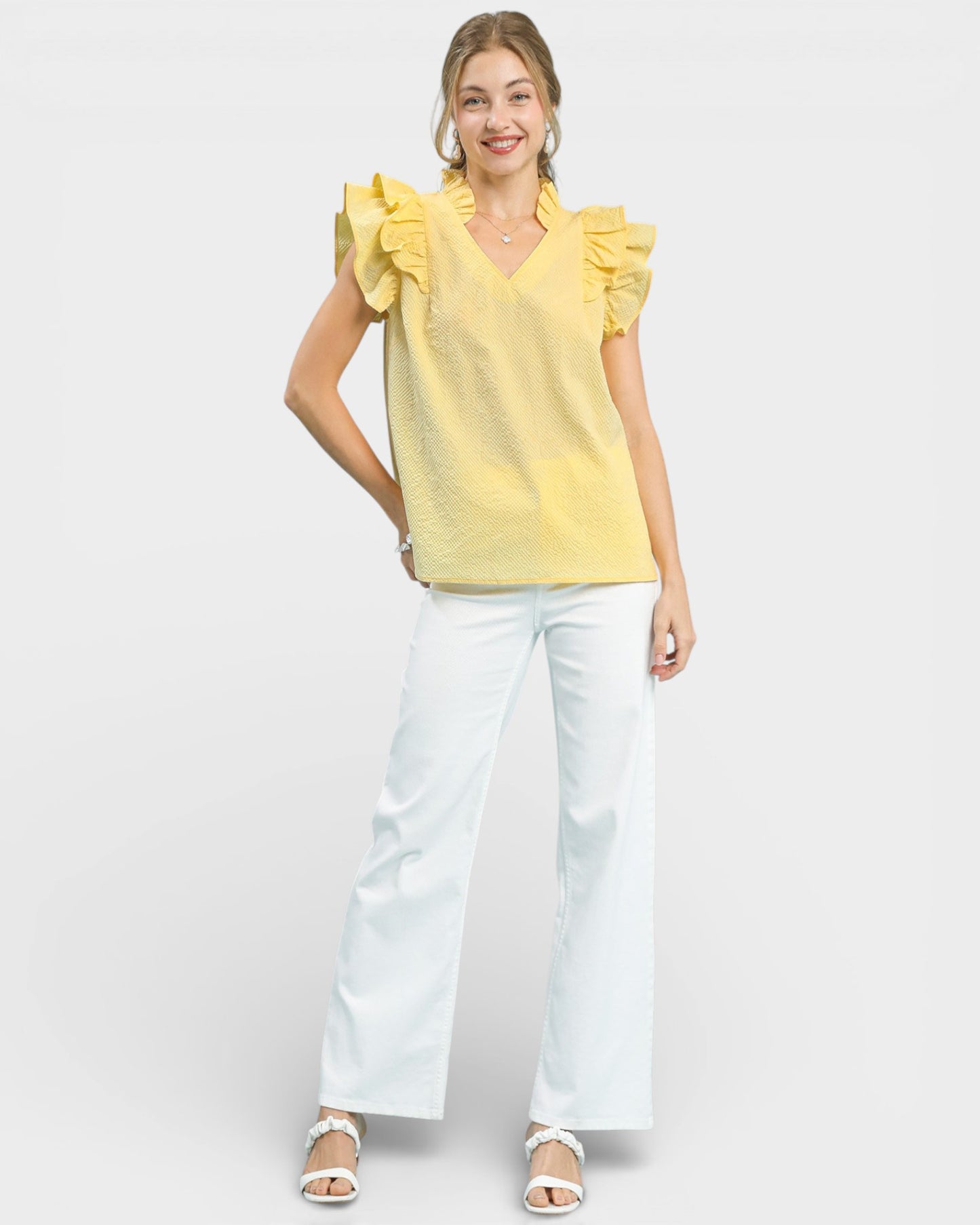 Woman wearing a yellow top with ruffled sleeves and white pants on a light gray background