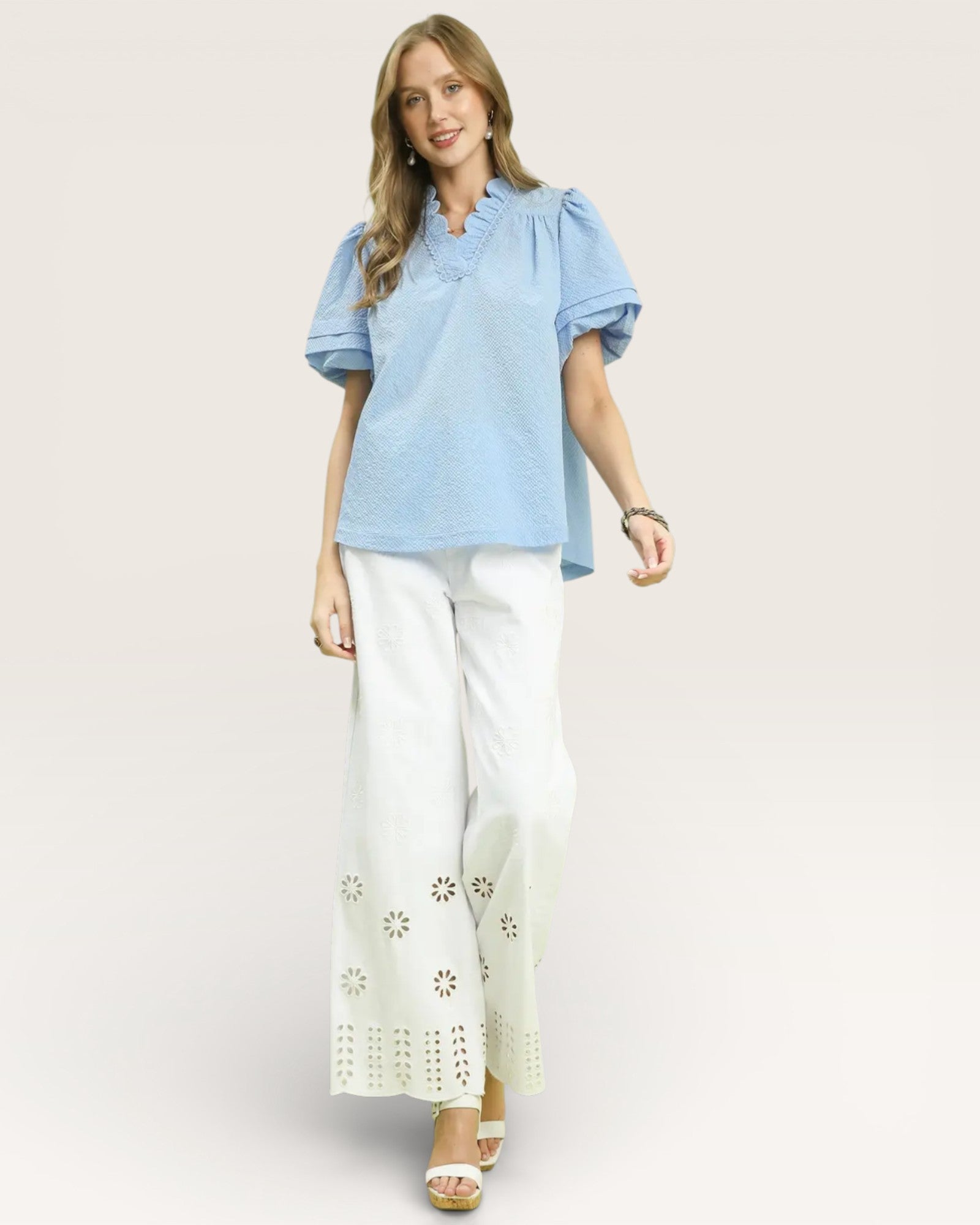 Woman wearing a light blue top and white pants with eyelet details on a plain background