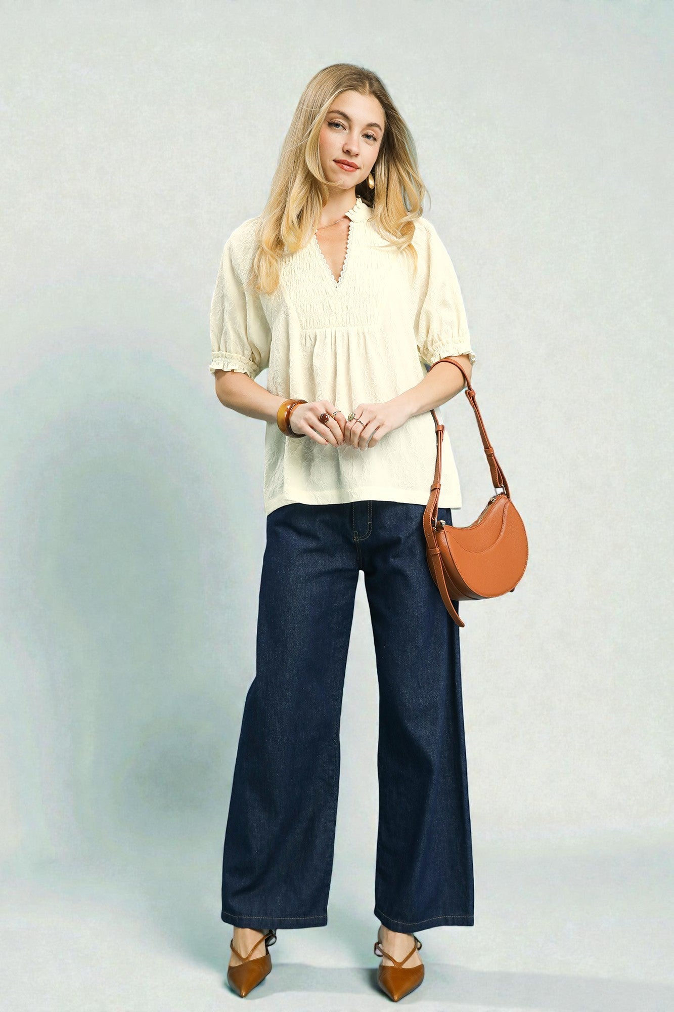 Woman wearing a cream blouse, navy pants, and brown shoes with a tan handbag on a light gray background